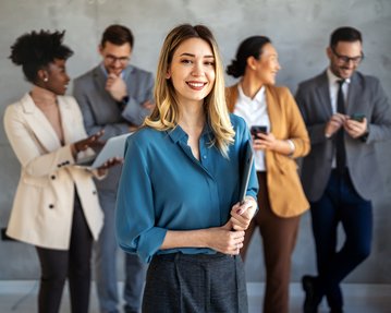 A smiling woman in a blue blouse is holding a binder, while four people in the background are communicating with each other and using devices.