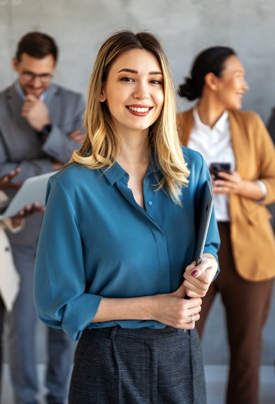 A smiling woman in a blue blouse is holding a binder, while four people in the background are communicating with each other and using devices.