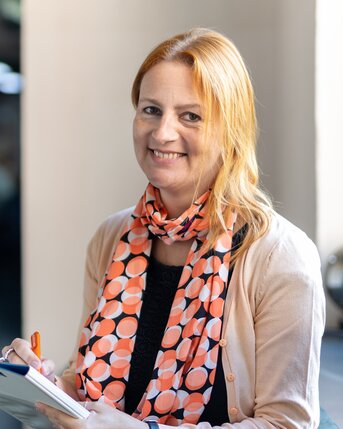 Eine lächelnde Frau mit langen, blonden Haaren trägt einen orangefarbenen Schal mit Punkten und hält ein Notizbuch in der Hand.