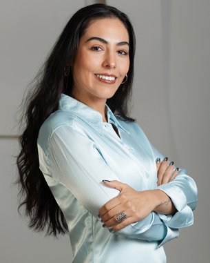 A woman with long, dark hair is wearing a light blue shirt and smiling as she poses with her arms crossed.
