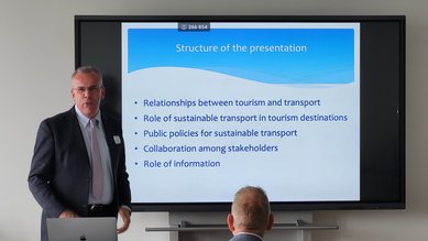 Ein Mann präsentiert vor einem Bildschirm mit einer Folie über die Struktur der Präsentation, die Themen wie Tourismus, nachhaltigen Transport und Zusammenarbeit behandelt. Im Vordergrund sitzt ein Zuhörer.