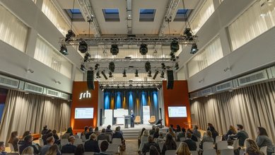 Ein Publikum sitzt in einem modernen Veranstaltungsraum, während ein Redner auf einer Bühne präsentiert. Beleuchtung und technische Ausstattung sind sichtbar.