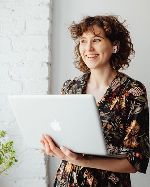 Eine lächelnde Frau mit lockigem Haar hält einen Laptop in der Hand. Sie trägt ein blumiges Oberteil und steht neben einer Pflanze in einem Topf. Der Hintergrund ist eine helle, gemauerte Wand.