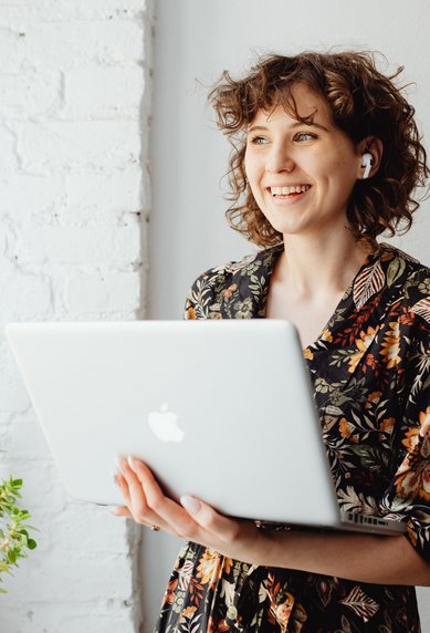 A smiling woman with curly hair is holding a laptop in her hand. She is wearing a floral top and standing next to a potted plant. The background features a bright, brick wall.