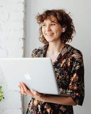 Eine lächelnde Frau mit lockigem Haar hält einen Laptop in der Hand. Sie trägt ein blumiges Oberteil und steht neben einer Pflanze in einem Topf. Der Hintergrund ist eine helle, gemauerte Wand.