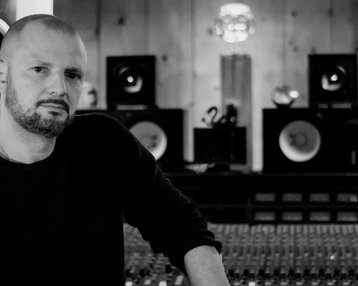 Mann mit Bart steht an einem Mischpult in einem Tonstudio, umgeben von Lautsprechern und einer Holzverkleidung.