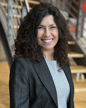 A woman with curly brown hair is wearing a gray sweater and a black blazer. She is smiling and standing in front of a staircase.