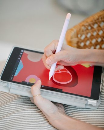 Eine Hand hält einen Stylus und zeichnet auf einem Tablet mit einem roten Hintergrund, der verschiedene bunte Formen zeigt. Die andere Hand stützt das Tablet auf einem gestreiften Kissen.