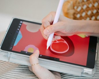 A hand holds a stylus and draws on a tablet with a red background displaying various colorful shapes. The other hand supports the tablet on a striped cushion.