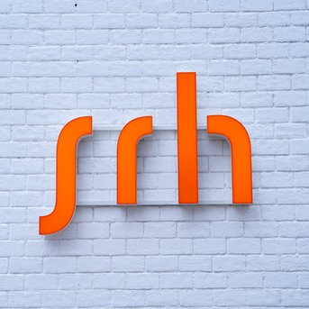 Ein orangefarbenes Logo mit den Buchstaben "srh" auf einer weißen Ziegelwand.