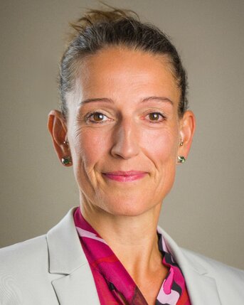 Eine Frau mit kurzen Haaren trägt einen blauen Blazer und ein pinkes Oberteil. Sie blickt direkt in die Kamera.