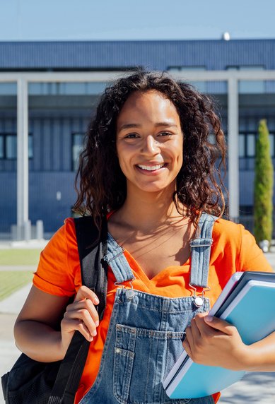 Eine junge Frau mit lockigem Haar trägt einen Rucksack und hält mehrere Bücher in der Hand, vor einem modernen Gebäude im Freien.