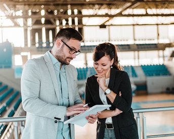 Ein Mann und eine Frau stehen in einem Sportstadion und betrachten gemeinsam ein Tablet. Beide scheinen an der Diskussion interessiert.