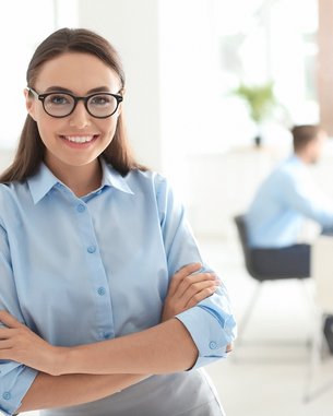 Eine Frau mit Brille und blauer Bluse steht lächelnd an einer Wand. Im Hintergrund sind mehrere Personen in einem Besprechungsraum zu sehen, die an einem Tisch sitzen und miteinander sprechen.
