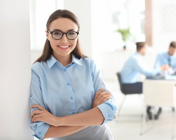 Eine Frau mit Brille und blauer Bluse steht lächelnd an einer Wand. Im Hintergrund sind mehrere Personen in einem Besprechungsraum zu sehen, die an einem Tisch sitzen und miteinander sprechen.