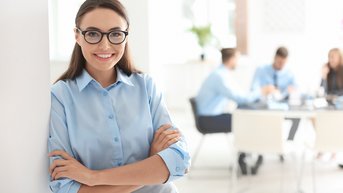Eine Frau mit Brille und blauer Bluse steht lächelnd an einer Wand. Im Hintergrund sind mehrere Personen in einem Besprechungsraum zu sehen, die an einem Tisch sitzen und miteinander sprechen.