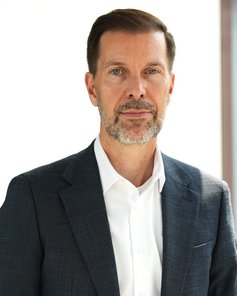 A man with short brown hair and a well-groomed beard is wearing a dark blue suit and a white shirt.