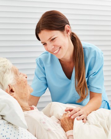 Eine Pflegekraft spricht freundlich mit einer älteren Patientin im Bett, während sie sich um sie kümmert.
