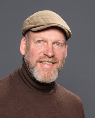 Man with a beard and a hat smiling at the camera. He is wearing a brown turtleneck sweater. The background is gray.