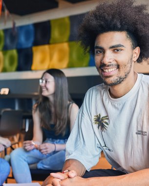 Ein junger Mann mit lockigem Haar sitzt lächelnd an einem Tisch. Im Hintergrund unterhalten sich zwei Frauen. Die Umgebung ist modern und freundlich gestaltet, mit bunten Wandpaneelen.