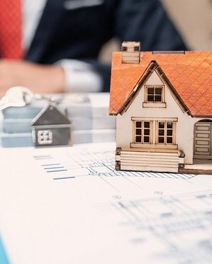 Ein Mann in einem Anzug schreibt an einem Tisch, während er eine kleine Modellhausfigur und einen Schlüssel auf einem Dokument betrachtet. Ein Laptop ist ebenfalls auf dem Tisch platziert.