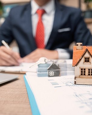 Ein Mann in einem Anzug schreibt an einem Tisch, während er eine kleine Modellhausfigur und einen Schlüssel auf einem Dokument betrachtet. Ein Laptop ist ebenfalls auf dem Tisch platziert.