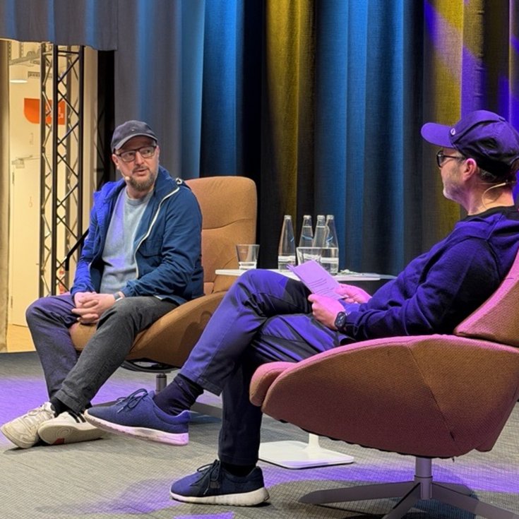 Two men are sitting in modern armchairs on a stage, engaged in conversation. In the background, colorful lights and curtains are visible.