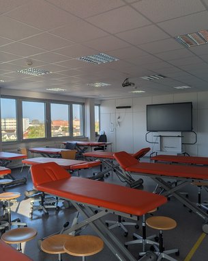 Ein heller Raum mit mehreren Behandlungsliegen in Orange, Stühlen und einem großen Bildschirm an der Wand. Fenster bieten Ausblick nach draußen.
