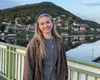 Junge Frau mit langen Haaren steht auf einer Brücke, trägt eine graue Strickjacke und lächelt, im Hintergrund eine ruhige Landschaft mit Hügeln und Häusern.