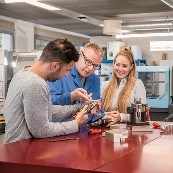 Three people in a modern workshop environment are examining a technical device together. A man is explaining something while a woman and another man listen attentively. In the background, machines are visible.