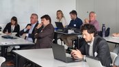 Eine Gruppe von Menschen sitzt an Tischen in einem Besprechungsraum. Einige schauen auf Laptops, während andere aufmerksam zuhören. Es gibt eine Tafel im Hintergrund.