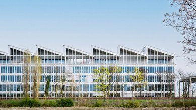 Modern office building with large windows and a distinctive, wave-shaped roof. Surrounded by trees and open landscape.