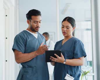 Zwei medizinische Fachkräfte in blauen Scrubs besprechen Informationen auf einem Tablet in einem modernen Krankenhausumfeld.