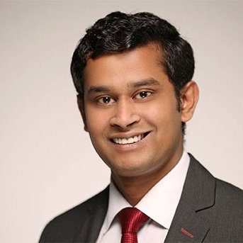 A young man with dark, curly hair is wearing a gray suit and a red tie. He is smiling warmly at the camera. The background is neutral and bright.