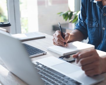 A person is writing in a notebook while working on a laptop. On the table, there is a smartphone and a coffee mug.