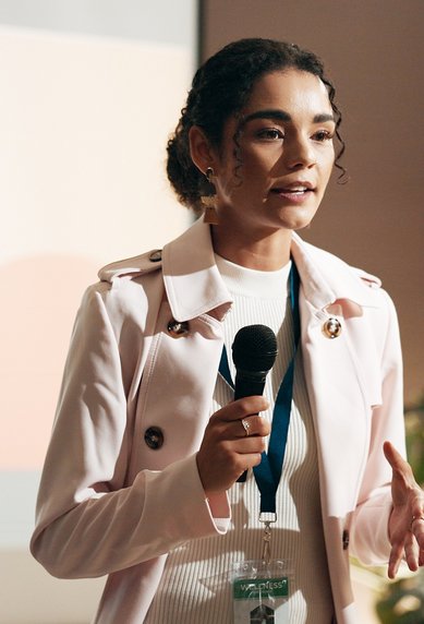 Eine junge Frau spricht mit einem Mikrofon auf einer Bühne, während eine Präsentation im Hintergrund sichtbar ist. Sie trägt einen weißen Blazer.
