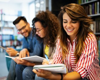 Drei Personen sitzen in einer Bibliothek, während eine Frau mit langen Haaren und gestreiftem Oberteil in ein Notizbuch schreibt. Zwei weitere Personen, ein Mann und eine Frau, schauen auf ihre Notizen und ein Tablet.