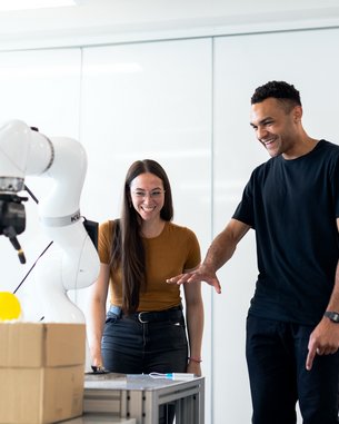 Zwei Personen stehen an einem Tisch und beobachten einen Roboterarm, der eine gelbe Kugel greift. Beide wirken interessiert und lächeln, während sie dem Roboter zuschauen.