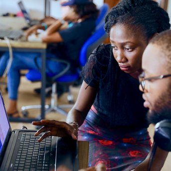 Two people are working on laptops in a modern office. One person is pointing at the screen while the other watches attentively. In the background, more people can be seen at their computers.
