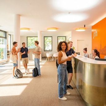 A group of young people is standing in a modern reception area while one person is working at the front desk. Some of them are holding drinks.