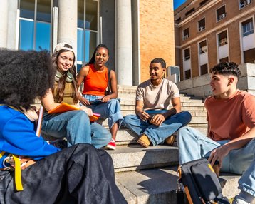 Five young adults are sitting on the steps in front of a building, discussing and sharing materials. They are dressed casually and appear relaxed.