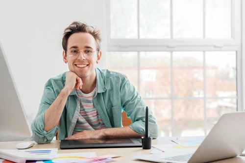 A young man with glasses sits at a desk, smiling, with a graphic tablet and various drawings in front of him.