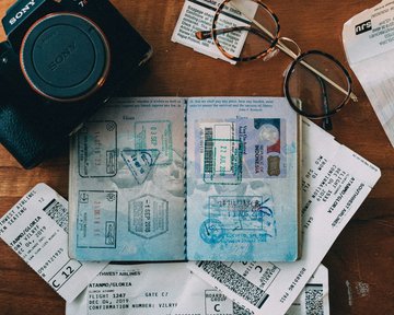 Passport with stamps, flight tickets, and a camera are laid out on a wooden table. A pair of glasses is also visible. The tickets display details about flights and gate information.