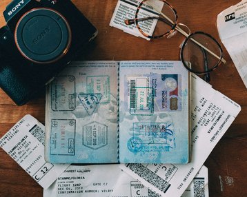 Passport with stamps, flight tickets, and a camera are laid out on a wooden table. A pair of glasses is also visible. The tickets display details about flights and gate information.