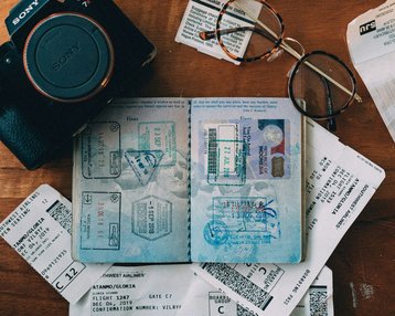 Passport with stamps, flight tickets, and a camera are laid out on a wooden table. A pair of glasses is also visible. The tickets display details about flights and gate information.