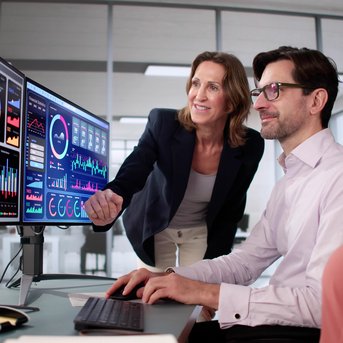 A woman is showing a man and a woman data visualizations and charts on several screens in a modern office.