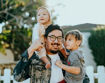 Ein Mann trägt ein Kleinkind auf den Schultern und steht neben einem weiteren Kind. Beide Kinder lächeln, während der Mann eine fröhliche Pose einnimmt. Im Hintergrund ist ein weißer Zaun und grüne Pflanzen sichtbar.