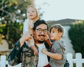 Ein Mann trägt ein Kleinkind auf den Schultern und steht neben einem weiteren Kind. Beide Kinder lächeln, während der Mann eine fröhliche Pose einnimmt. Im Hintergrund ist ein weißer Zaun und grüne Pflanzen sichtbar.