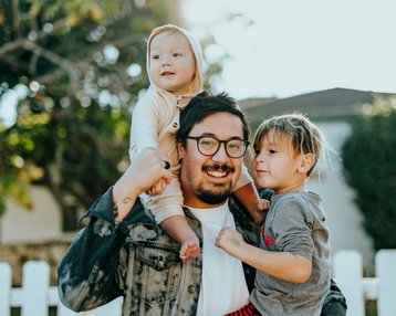 Ein Mann trägt ein Kleinkind auf den Schultern und steht neben einem weiteren Kind. Beide Kinder lächeln, während der Mann eine fröhliche Pose einnimmt. Im Hintergrund ist ein weißer Zaun und grüne Pflanzen sichtbar.