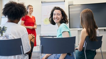 A group of four people is sitting in a seminar room while a woman in a red dress is giving a presentation.